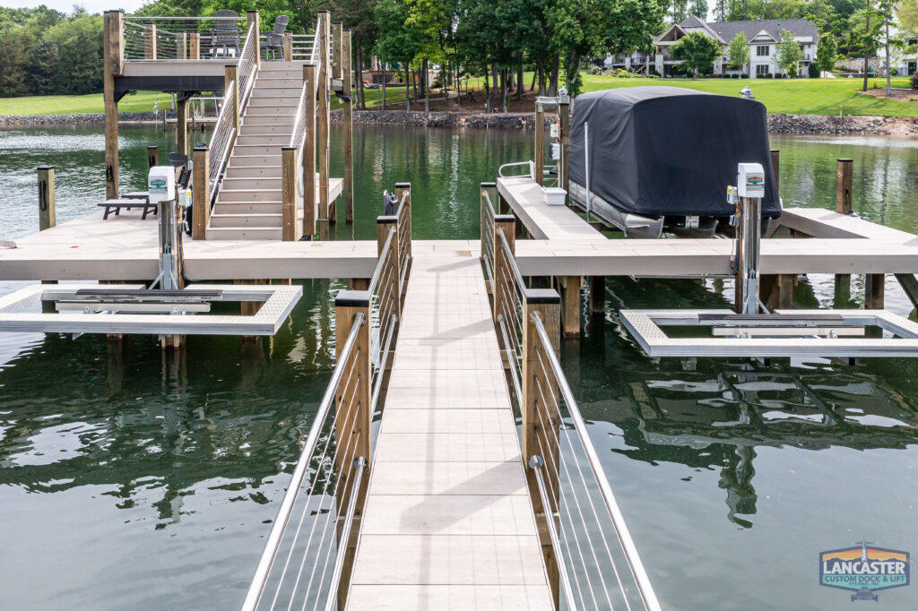 A wooden dock extends over a lake with multiple boat lifts, some covered, designed for easy access. The dock features handrails and a staircase leading down to the water. Trees and houses are visible in the background across the lake. A "Lancaster" sign is present.