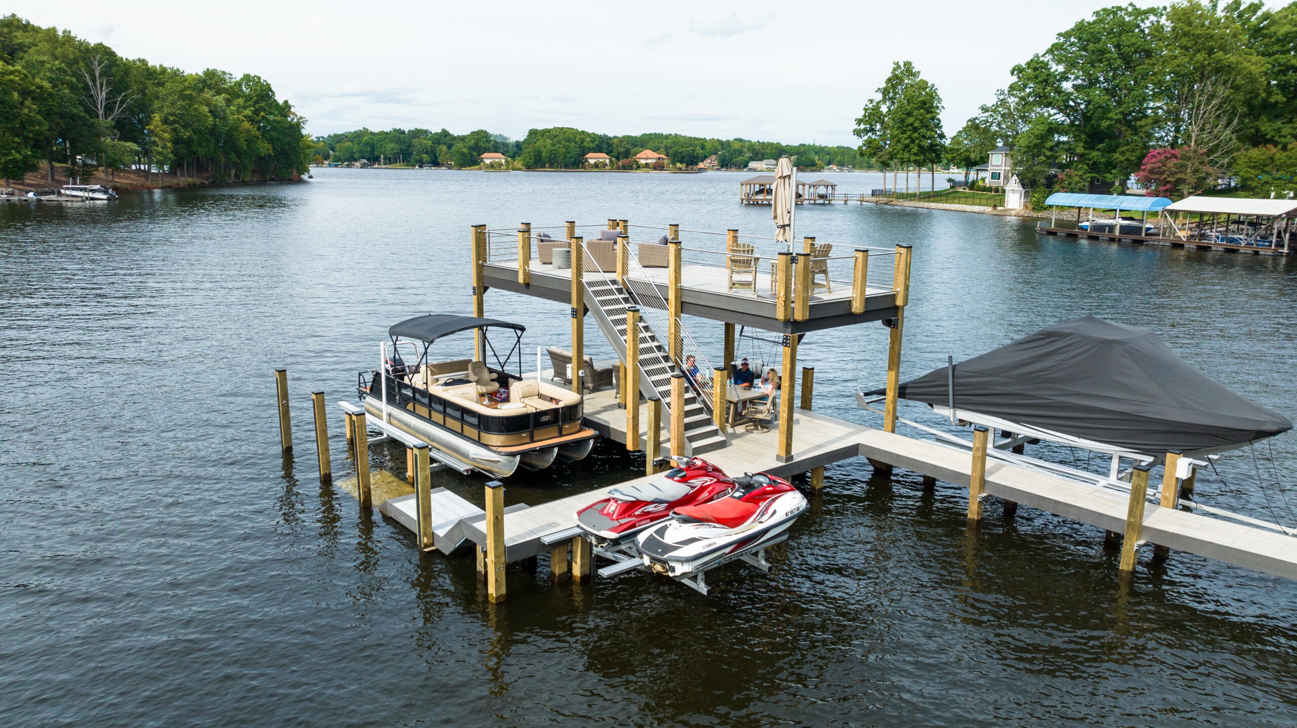 A dock structure extends into a lake, featuring a boat and two jet skis. A staircase leads to an elevated deck above. Several people are on the dock and deck, surrounded by lush greenery and a few houses in the background across the water.