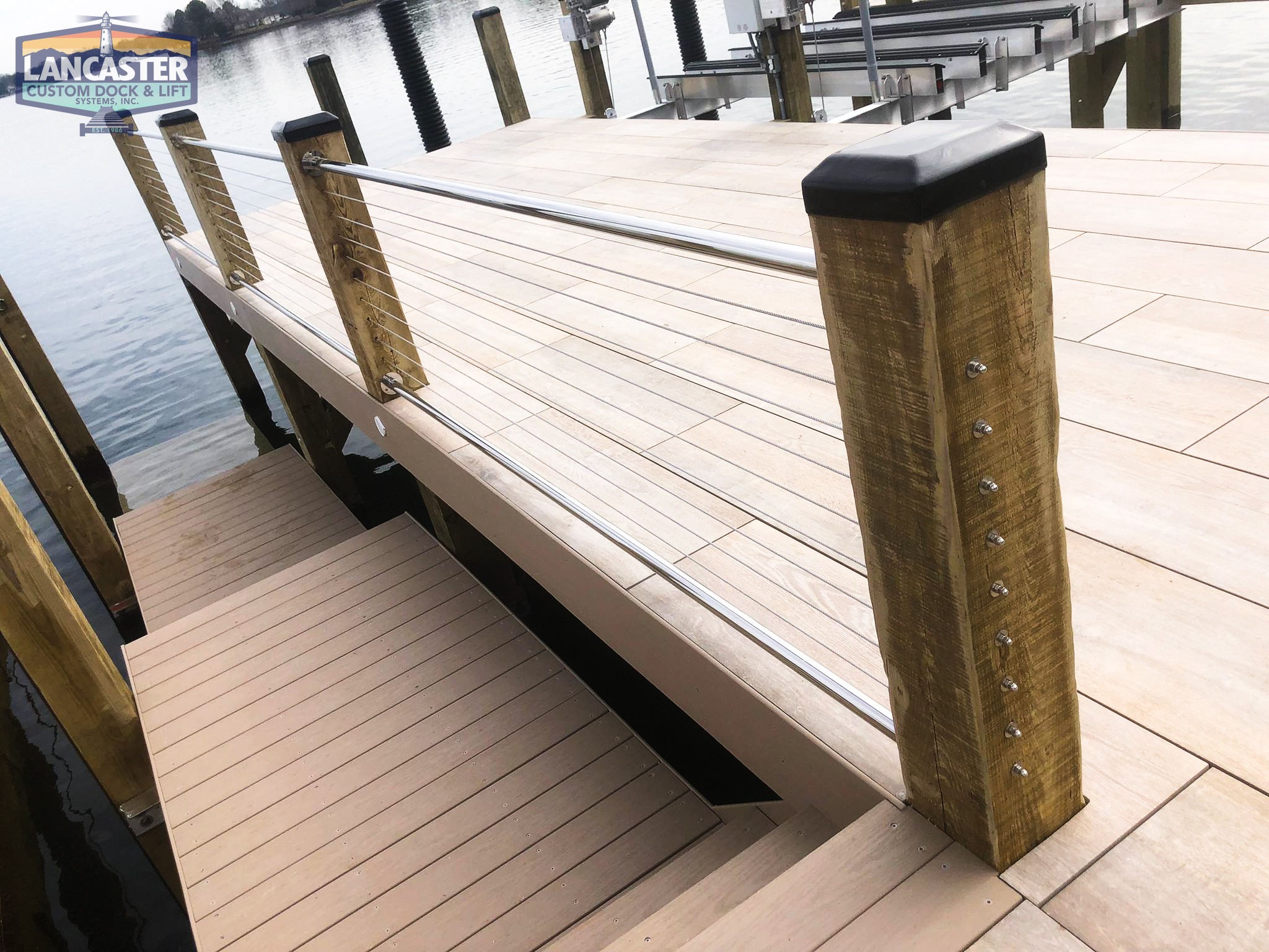 An elevated wooden dock with a cable railing system overlooking a calm body of water. The dock features sturdy wooden posts with metal cables and black caps on the posts for added support. A "Lancaster Custom Dock & Lift" sign is visible in the upper left corner.