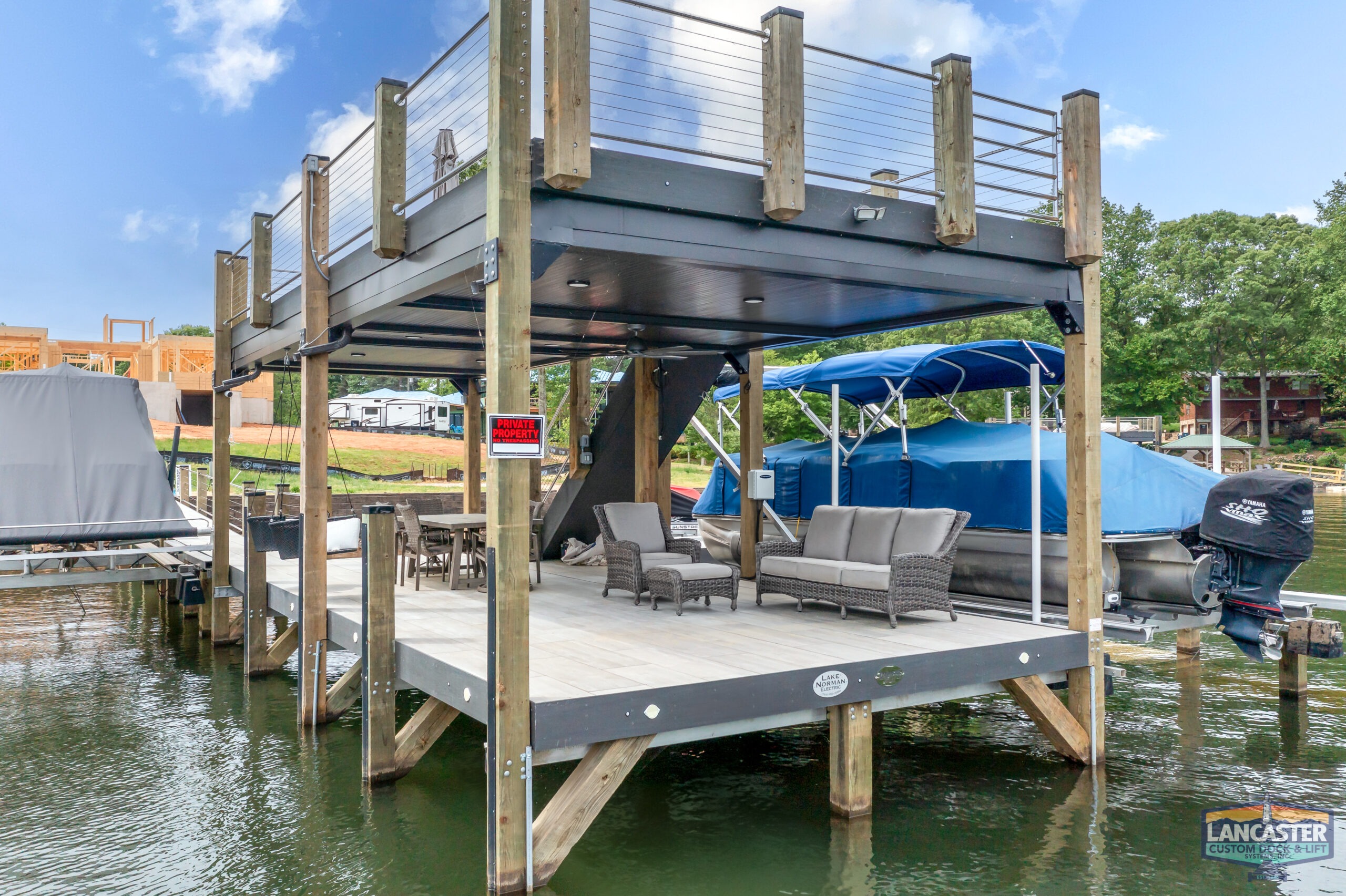 A covered boat dock with wooden pillars features outdoor furniture, including a sofa and chairs. The dock has an upper deck surrounded by cable railings. Several boats are visible, and a "Lancaster Custom Docks" sign is affixed to a pillar.