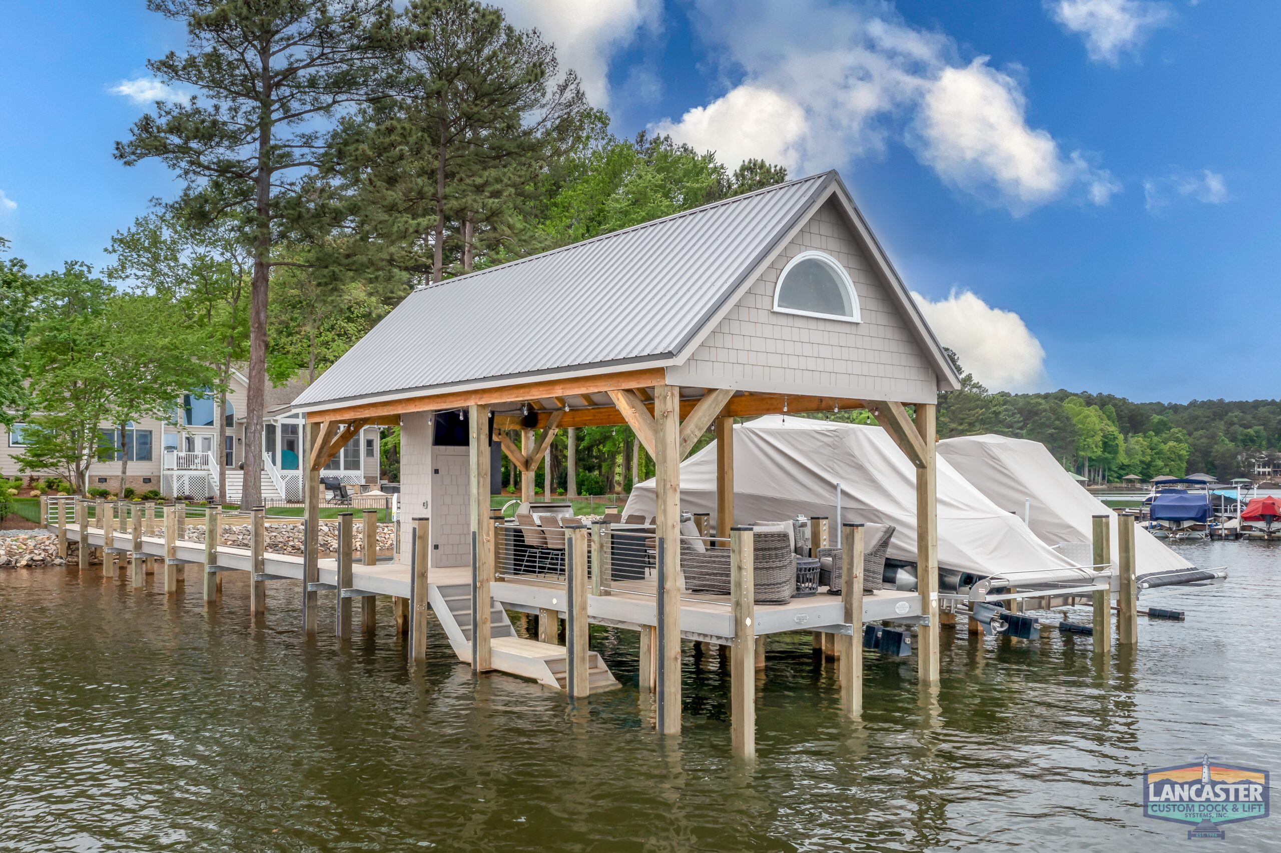 A modern boat house on a lake with a tin roof, surrounded by a wooden dock and greenery. Several covered boats are docked next to the house. The Lancaster custom dock sign is in the bottom right corner of the image. Blue sky with some clouds above.