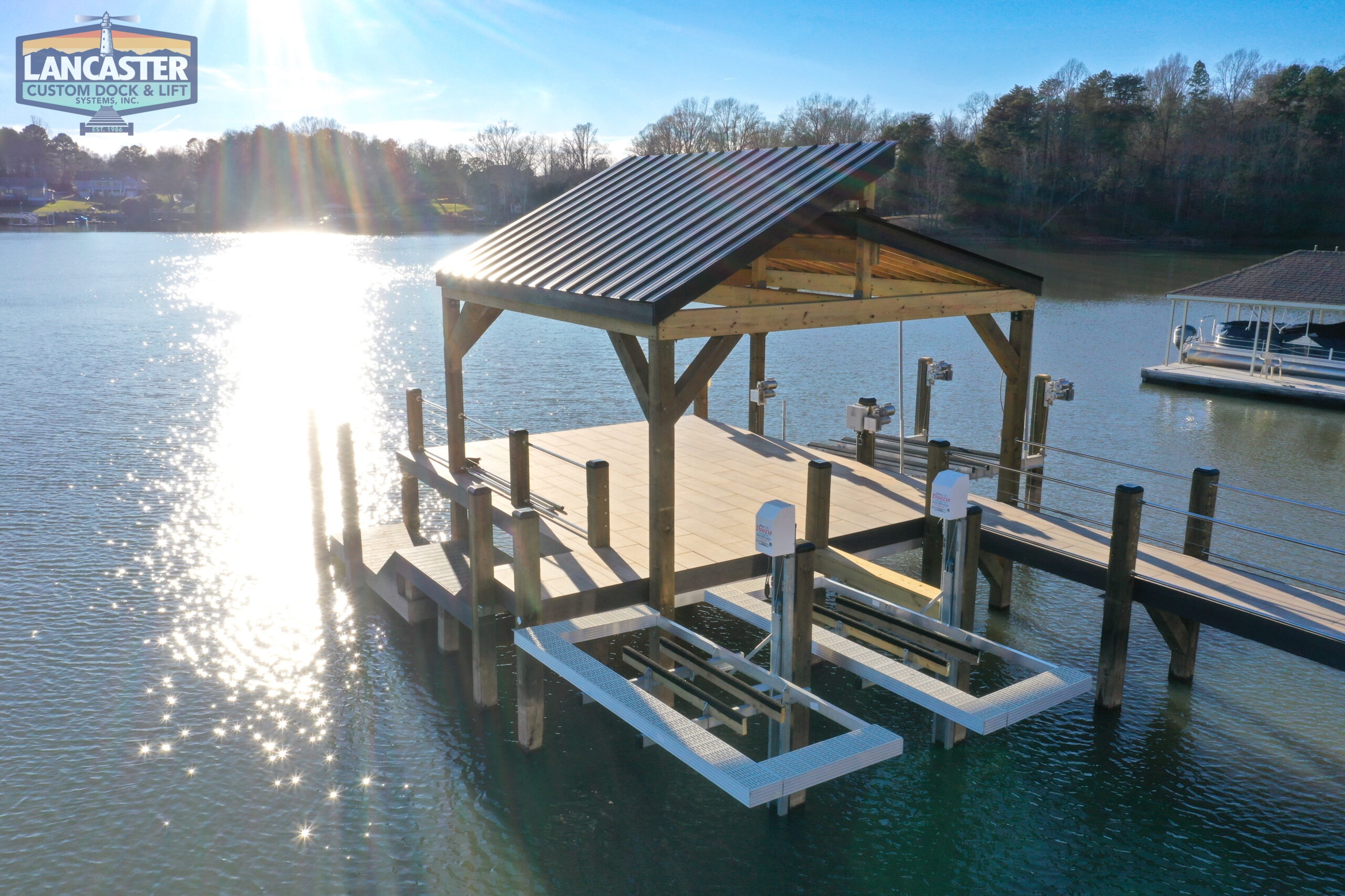 A wooden dock with a metal-roofed shelter extends over a serene lake, with sunlight reflecting off the water. Two boat lifts are attached to the dock. A company logo for Lancaster Custom Dock & Lift is visible in the upper left corner. Trees line the distant shore.