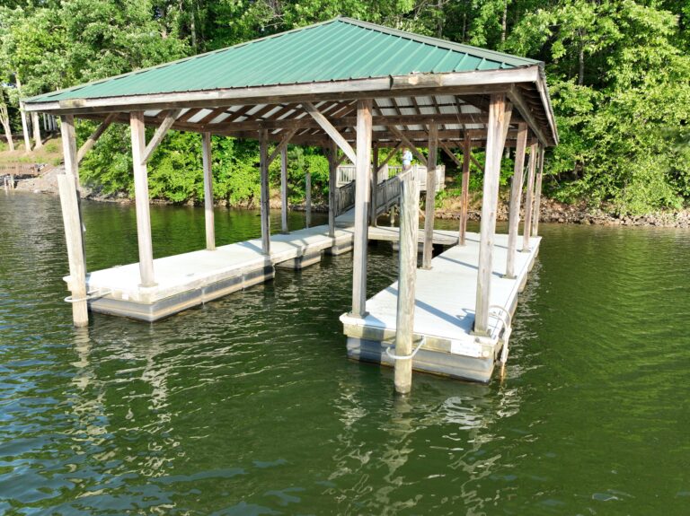 A wooden boat dock with a green metal roof extends over a calm body of water. It features two covered slips and is surrounded by lush green trees on the shoreline.