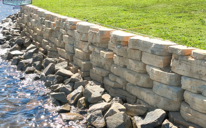 A well-constructed retaining wall made of large, beige stone blocks with jagged edges stands by a water body. Below the wall, clusters of smaller rocks are scattered. A green lawn stretches above the wall, and a sign reads "Lancaster Custom Dock & Lift".