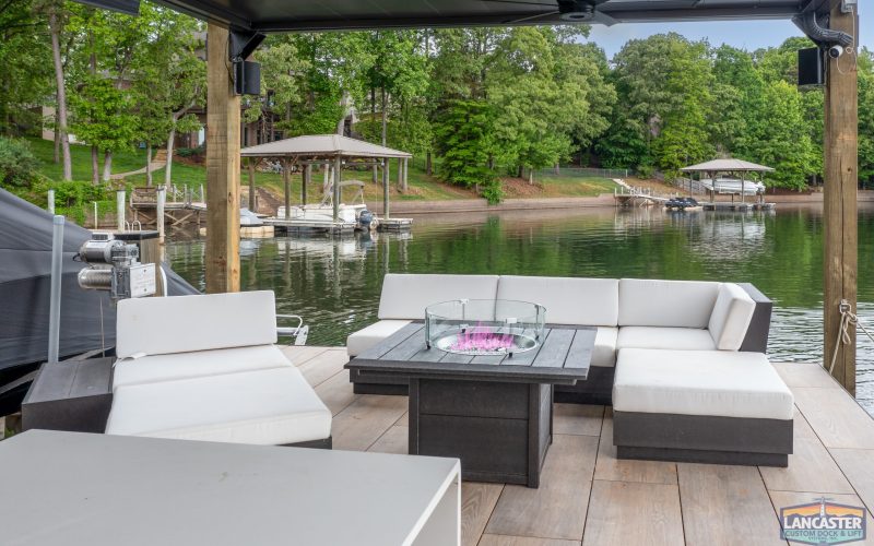 A luxurious outdoor seating area with modern white cushioned furniture and a square fire pit table sits on a wooden dock overlooking a serene lake with several docks and boats nearby. Trees line the shore, and the atmosphere is calm and inviting.